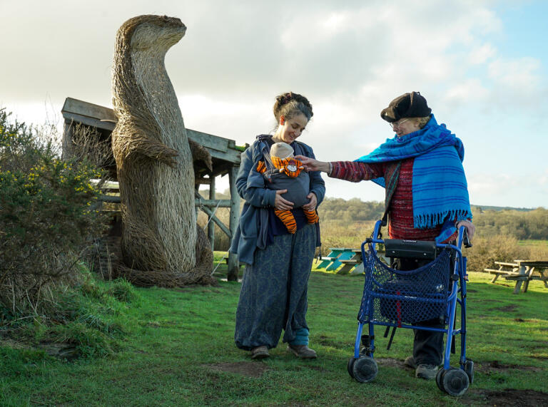 Eine Frau mit einem Baby und eine Frau mit einem Rollator vor der Weidenskulptur eines Tieres.