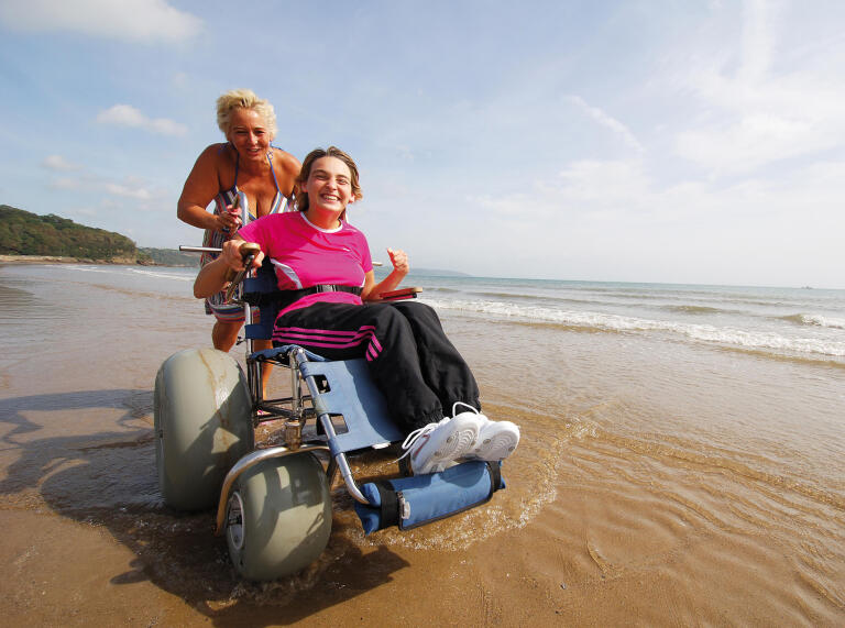 A lady using a beach wheelchair being pushed by another lady on a beach.