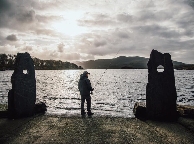man stood fishing by lake between two stone pillars.