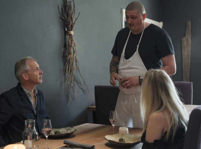 chef stood talking to man and woman sat at table.