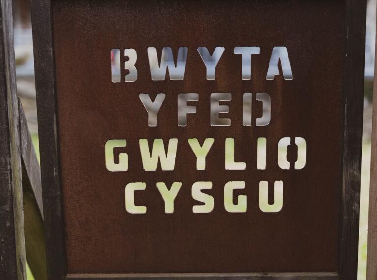 A metal sign with the Welsh words Bwyta, Yfed, Gwylio Cymsu - which translate to Eat, Drink, Watch, Sleep