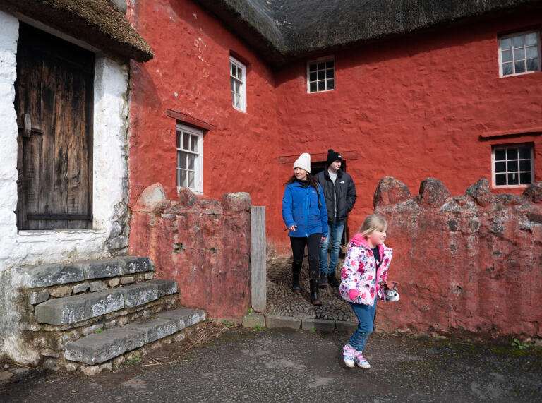 An adult and two children outside a red painted farmhouse.