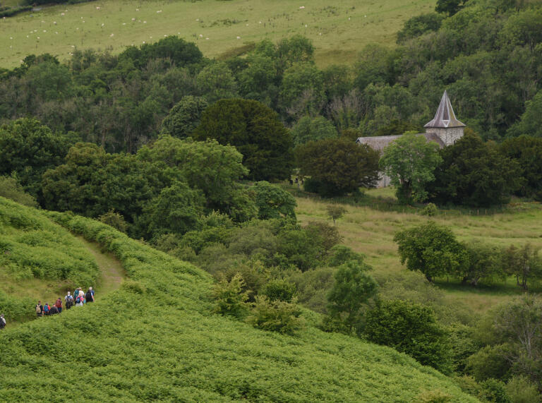 A  group of walkers on a grassy hillside path with a grey stone church in the distance.