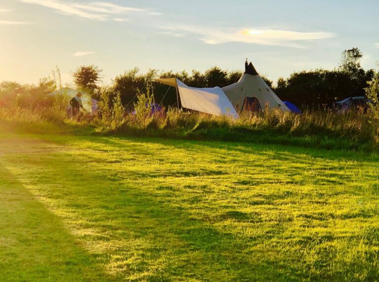 Tipi in a field full of green grass. The sun shines as the sun starts to sunset.