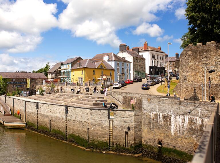 A river quayside with castle walls and colourful buildings.