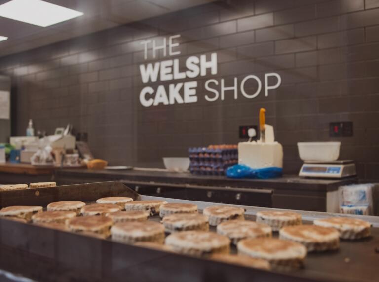 A welsh cake stall at a market with The Welsh Cake Shop sign in the background. Welsh cakes are being cooked on the griddle in the foreground. 