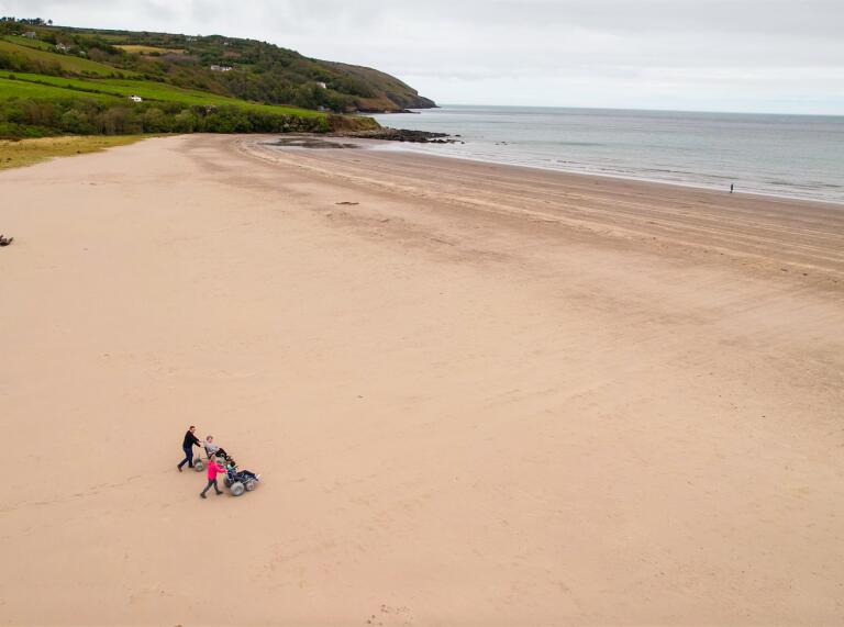 A drone shot of a wide sandy beach with two people using beach wheelchairs.
