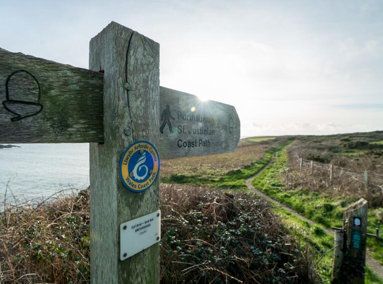A wooden Wales Coast Path signpost pointing along a grassy cliff top pathway.