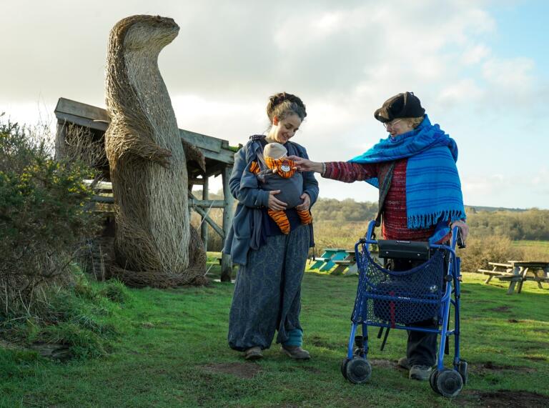 A woman with a baby and a woman with a walking frame in front of a willow animal.