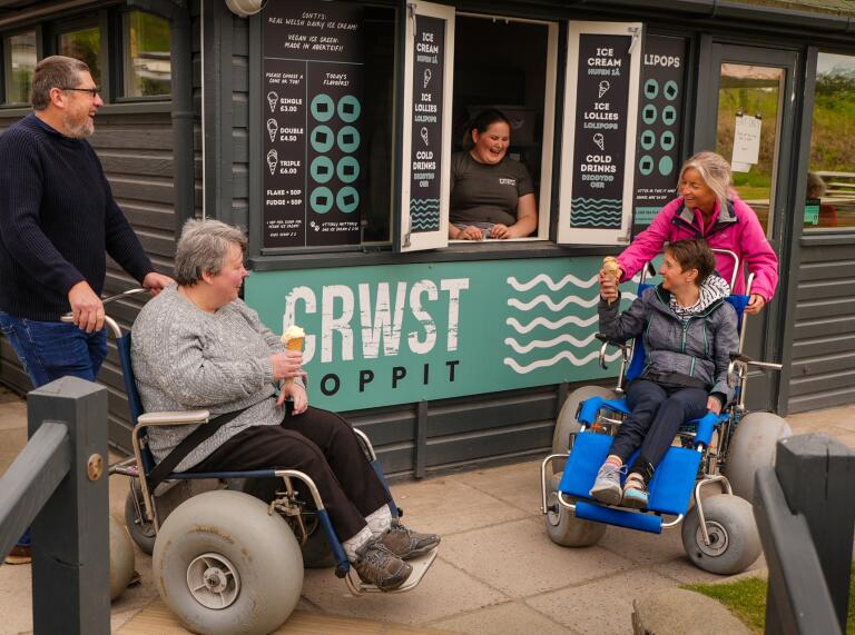 Two ladies using beach wheelchairs eating ice creams outside a cafe.