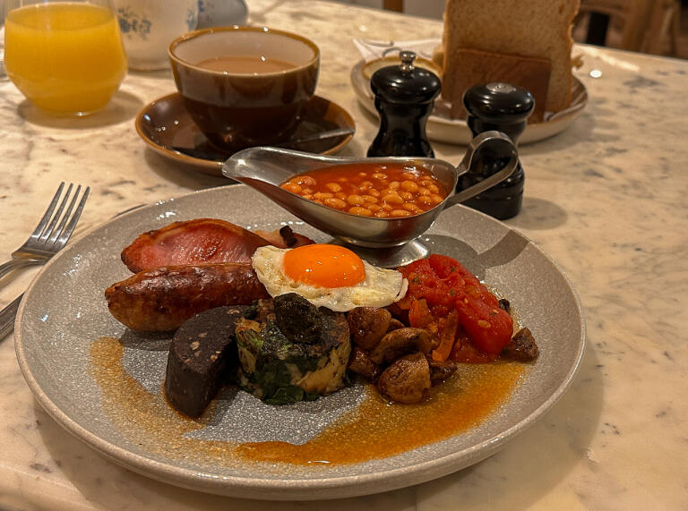 A white plate full of sausage, bacon,  laverbread, fried egg, black pudding, baked beans, tomatoes, mushrooms and coffee and juice on the side.