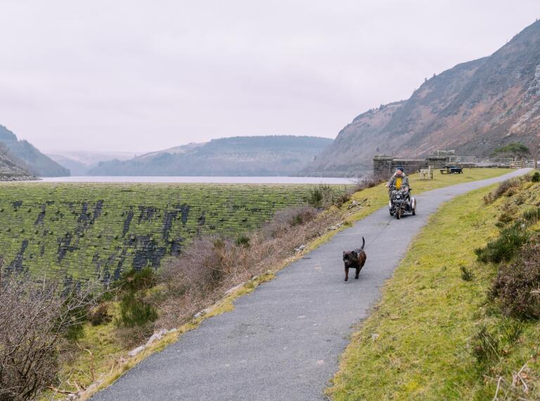 A lady on an electric buggy and her dog on a wide path next to a dam and reservoir.