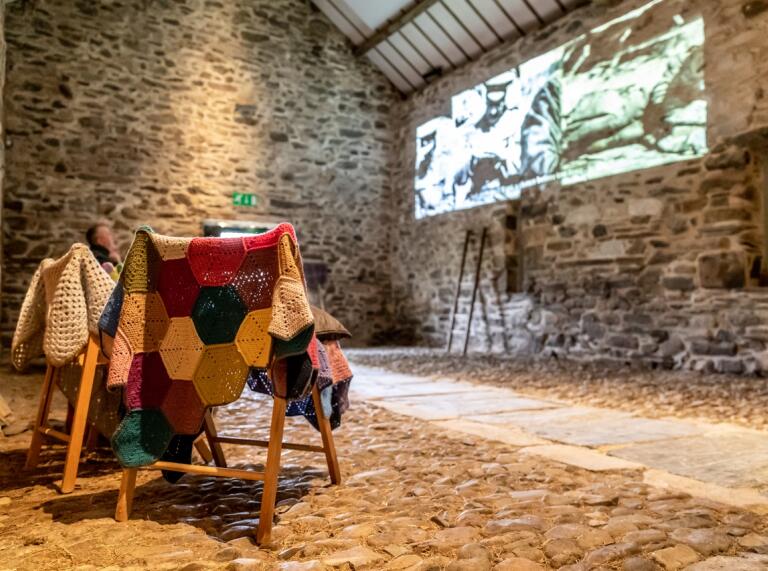 Inside a stone-walled cowshed restored into an interpretation centre. A film is being projected onto the wall and colourful crochet blankets are draped over wooden chairs.   