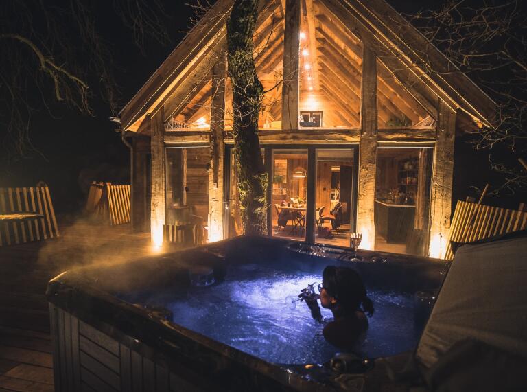 Night time. A lady sipping a glass of probably wine in a hot tub on a treehouse decking.