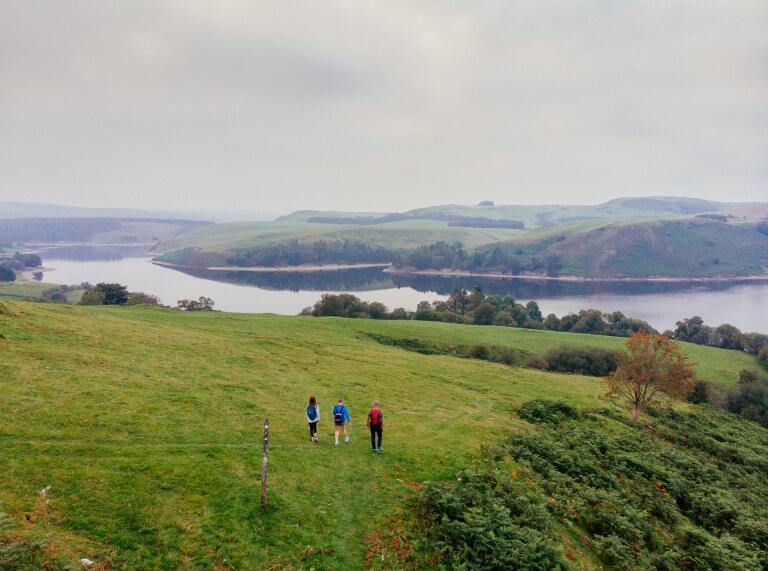 Three people walking along a footpath though a field, overlooking a wide reservoir.