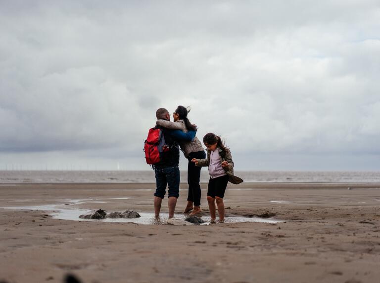 Two adults and a child paddling in a rockpool on a sandy beach.