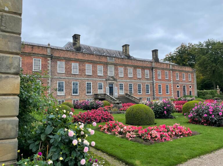 A large red brick stately home with colourful flower borders in front. 