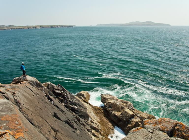 Ein Mann der auf einem Felsen hoch über dem Meer steht und einer Insel im Hintergrund.