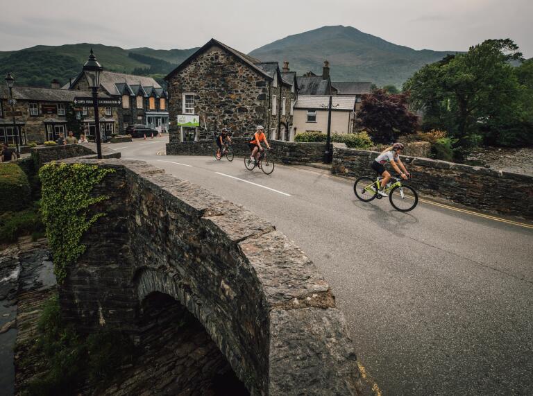 Three cyclists riding over a stone river bridge in a pretty village.