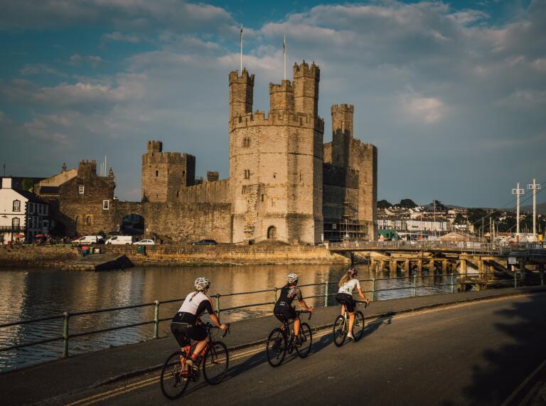 Three road cyclists on a road in front of an imposing castle. 