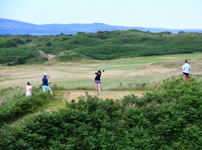 A female golfer taking a shot on a golf course surrounded by grassy dunes.