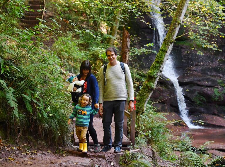 Two adults and two small children on a path next to a waterfall in woodland.