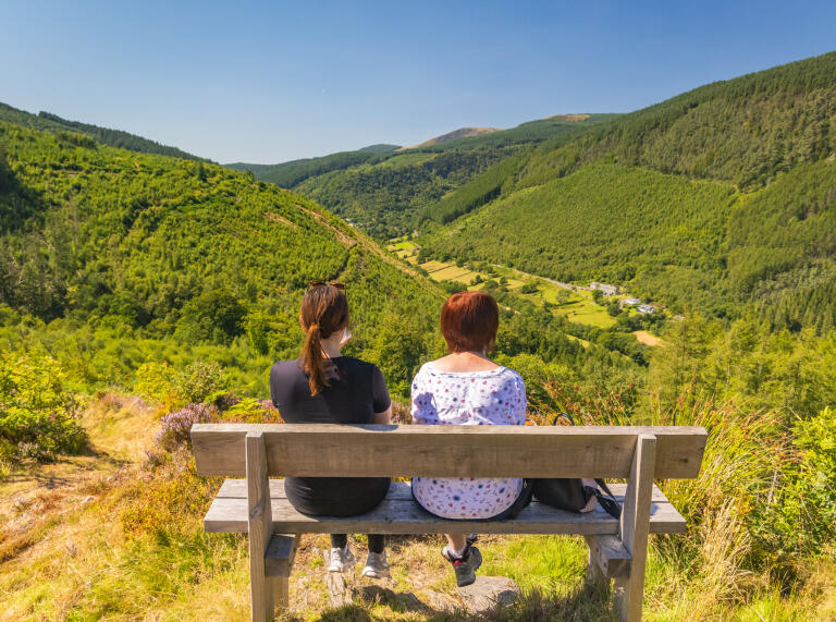 Two women sat on a bench overlooking a woodland sided valley and a small hamlet.