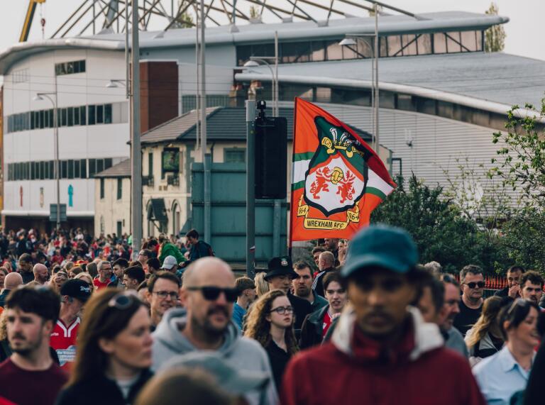 Crowds of people on a street outside a football stadium, with a 'Wrexham AFC' banner held high.