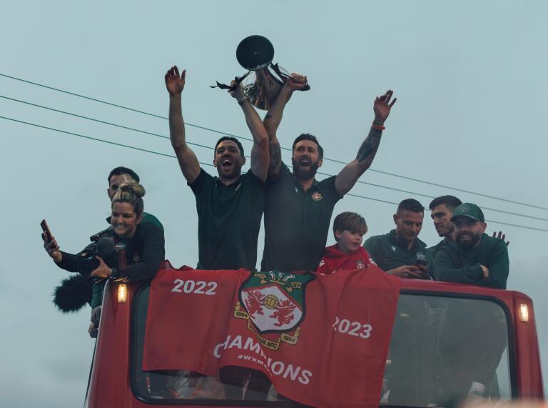 An open top bus with several people on the top deck. Two people are holding a trophy above their heads.
