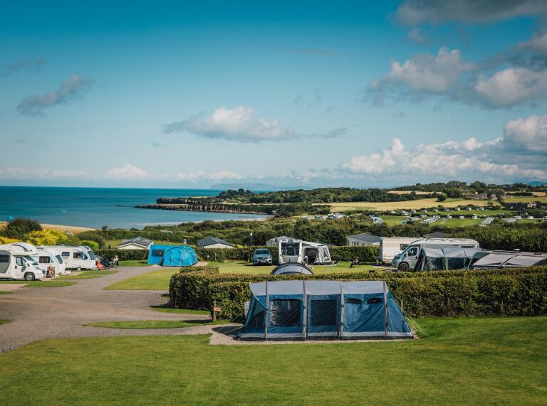 Campsite with tents and caravans pitched in fields, separated by hedges. The sea can be seen in the distance.