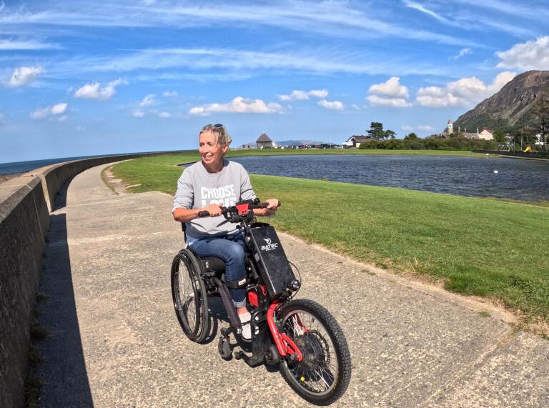 person  sat on a motorised trike, on a concrete coastal path with grass to the left and a blue sky above.