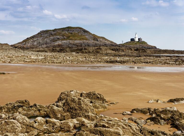 A sandy beach with rockpools and cliffs, with a lighthouse in the distance.