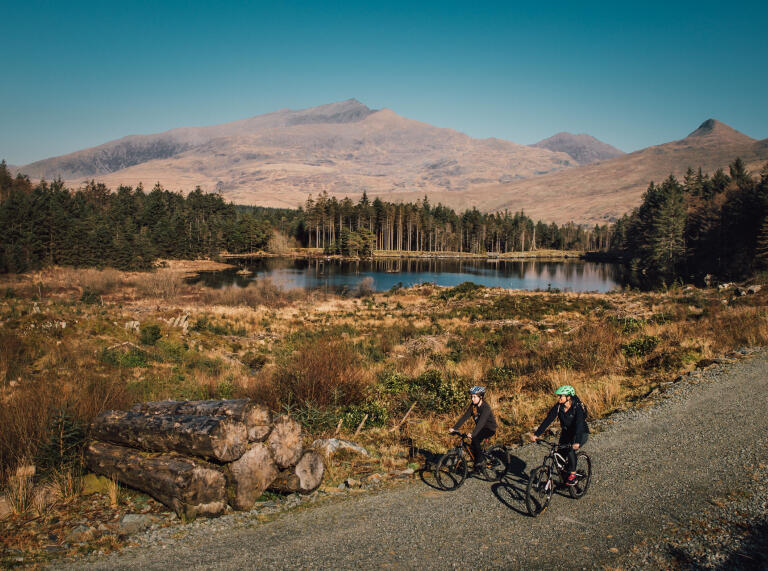 Two people riding mountain bikes on a gravel path by a lake.