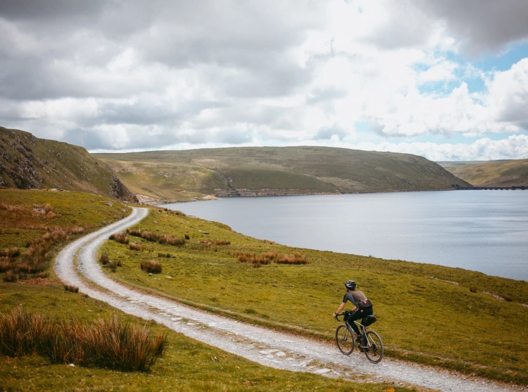 A cyclist riding along a gravel track alongside a mountain reservoir.