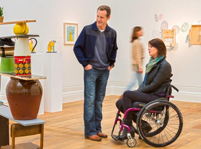 Woman using wheelchair and a standing man at an art gallery.