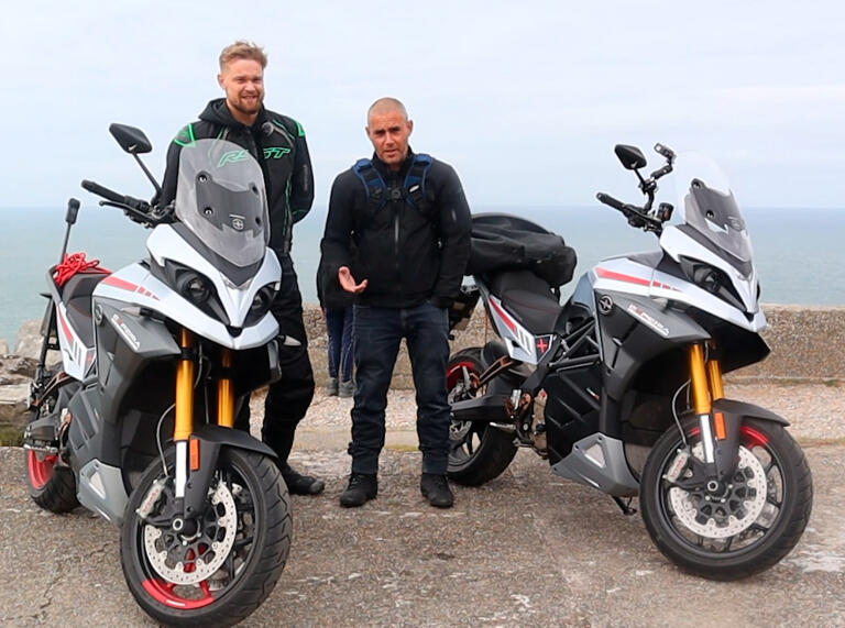 Two men stood by parked motorbikes with the sea in the background.