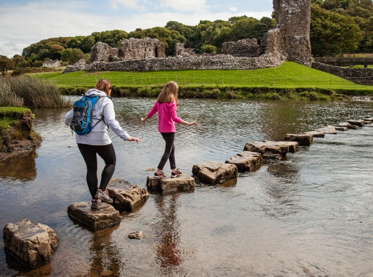 two young females walking on stepping stones with castle ruins in the background.