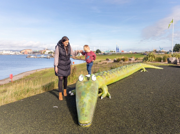 Boy standing on large crocodile-shaped bench, with a woman holding his hand.