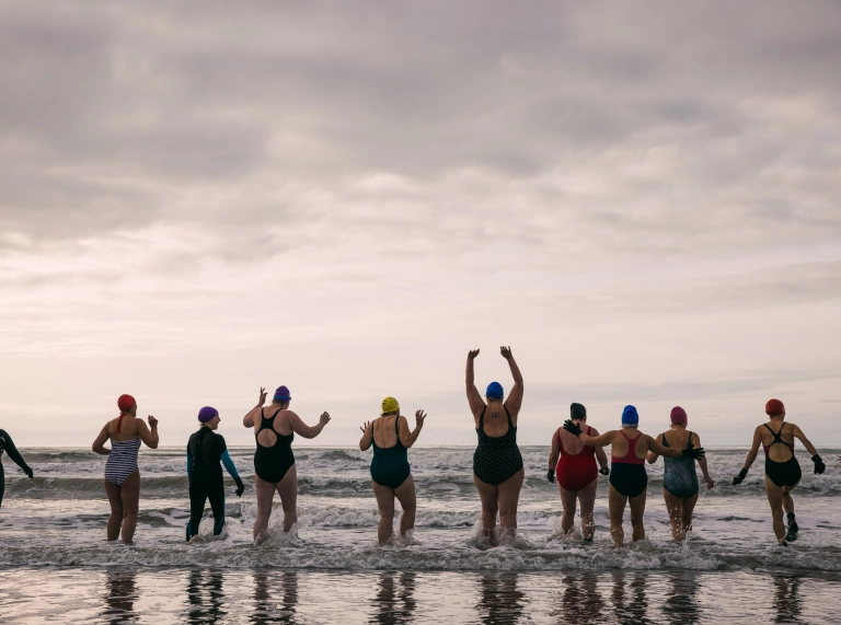 ten individuals going cold water swimming wearing swimsuits and swim caps. Seen from behind, they are stood in the shallow waves of the sea. One person is holding their hands above their head, others have their hands in the air.