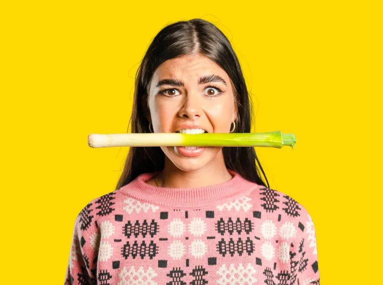 A woman wearing a pink jumper with a leek in her mouth.