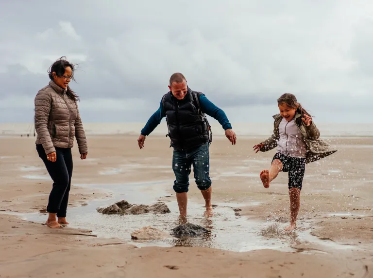 Two adults and a child splashing about in a rockpool on a sandy beach.
