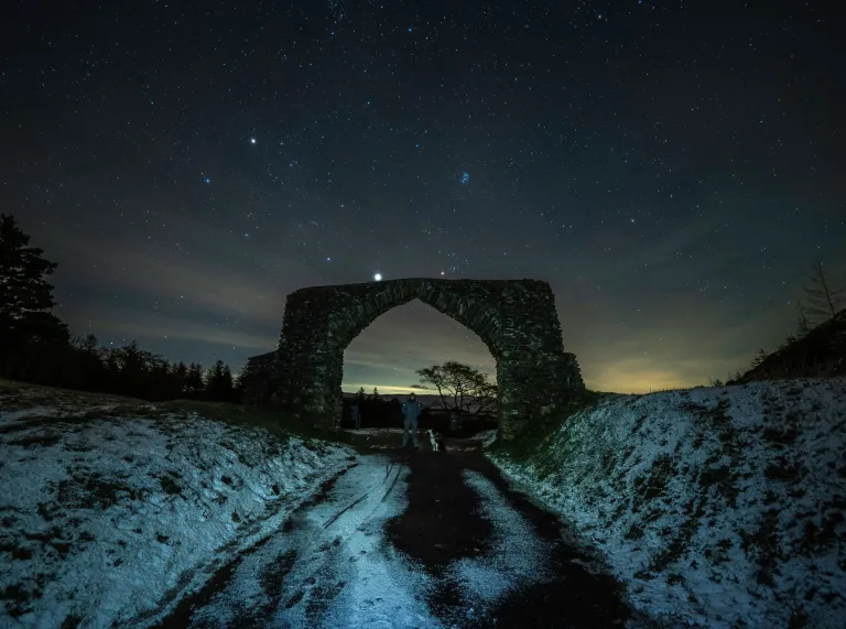 A stone built archway over a snowy narrow road at night. The stars are bright in thedark skies.