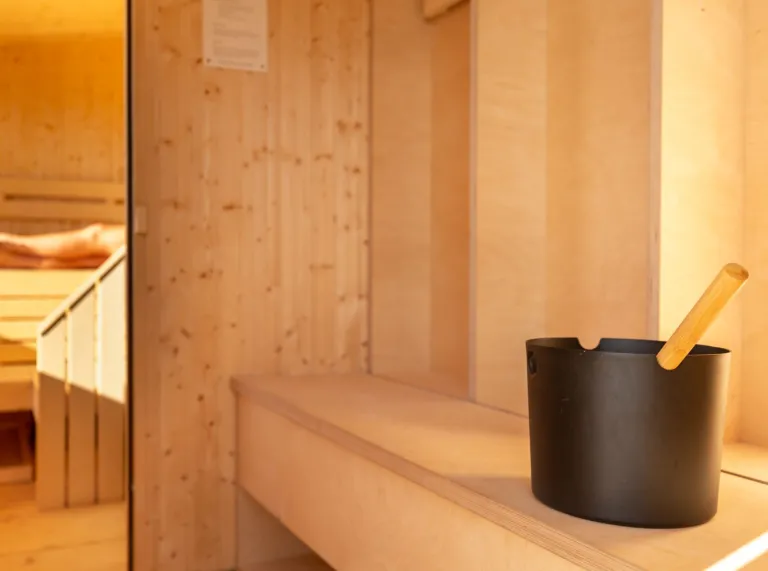 The inside of a wooden sauna with wooden walls and bench with a black bucket with wooden utensil in it.