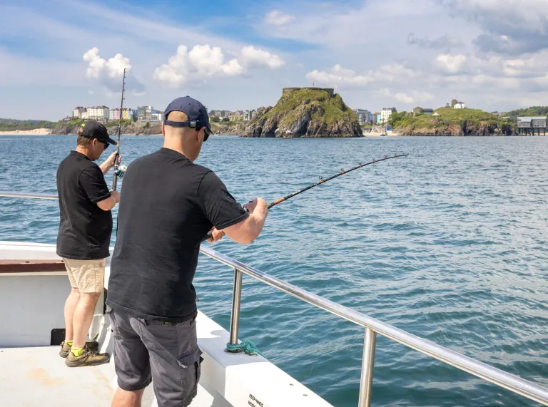 two men fishing from a boat on a sunny day with Tenby in the distance.