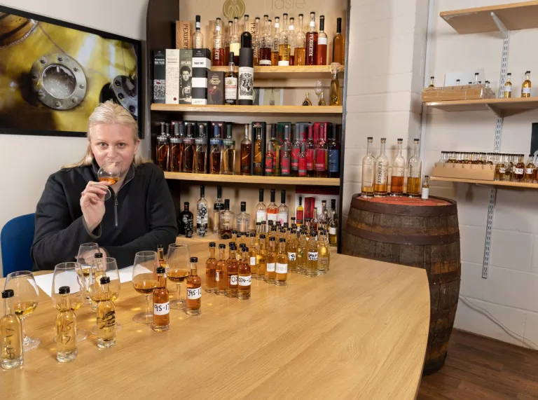 woman with glass of whisky with bottles and glasses on table and shelf.