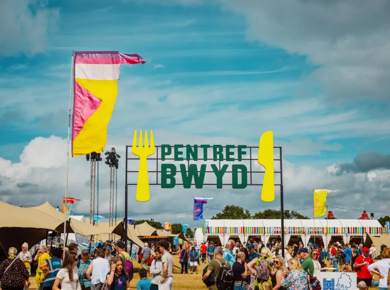 Sign in Welsh reading 'pentre bwyd' with large fork and knife, crowds of people and food tents.