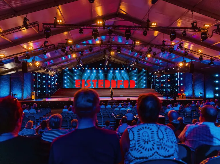 People watching performer on stage with large neon sign 'Eisteddfod'.