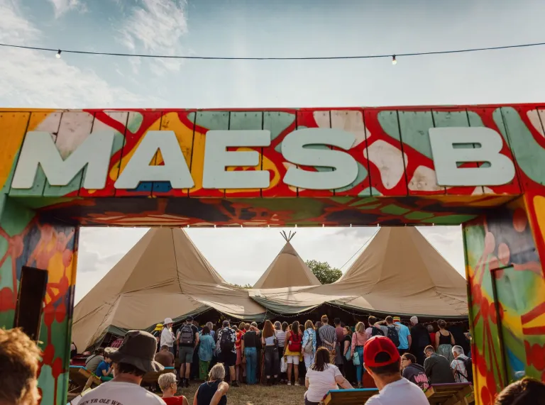 Sign reading 'Maes b' with festival tents and people.