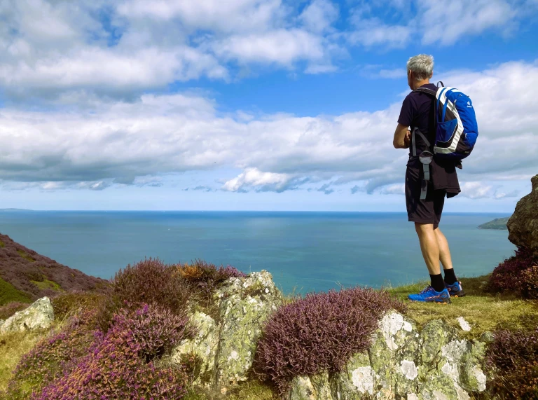 Mann mit blauem Rucksack, der auf einem Felsen steht und aufs Meer blickt.