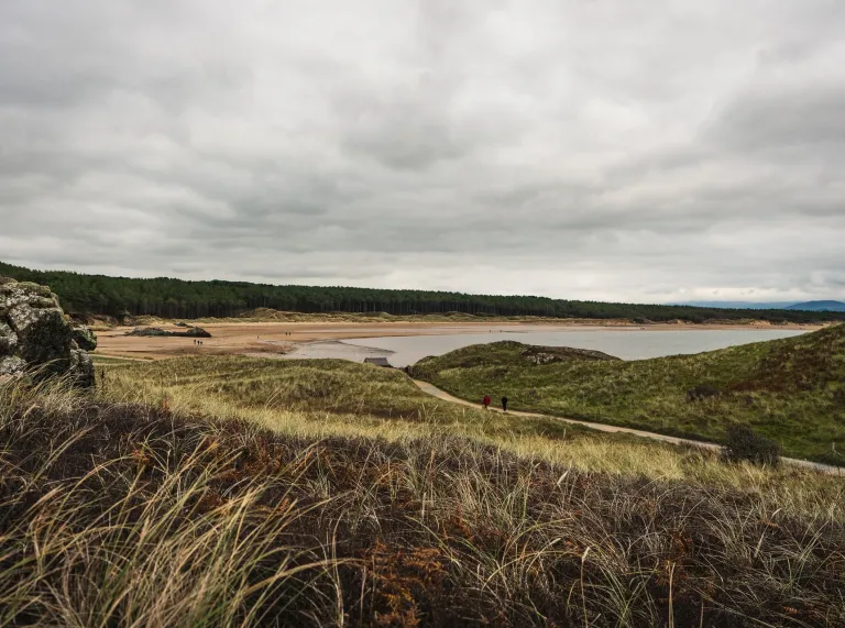 View of a sandy beach and tidal bay framed by grassy dunes and rocky outcrops under a cloudy sky.